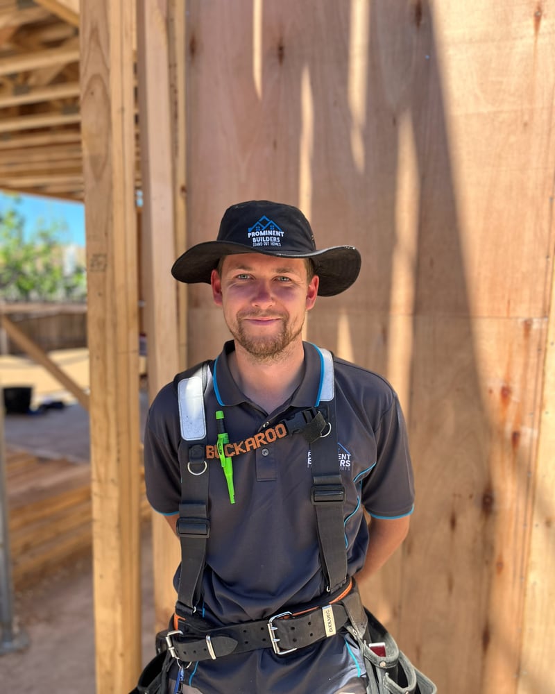 Bailey, carpenter at Prominent Builders, in a wide-brim hat and tool harness in front of a timber frame