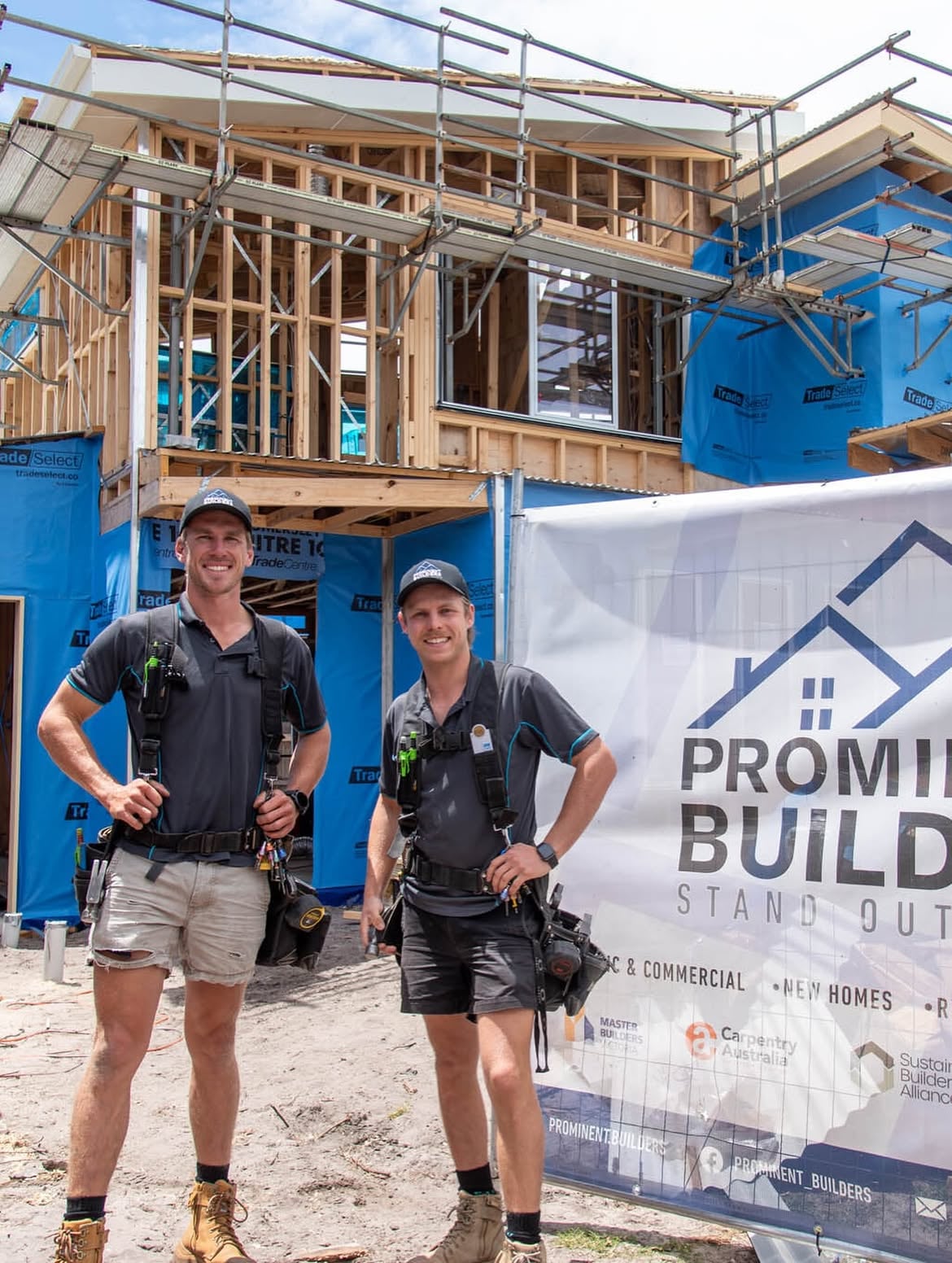 Joshua Lambert and Nathanael Furlong on site in Prominent Builders uniform and tool harnesses, in front of a two-storey timber-framed home under scaffolding, beside a Prominent Builders banner