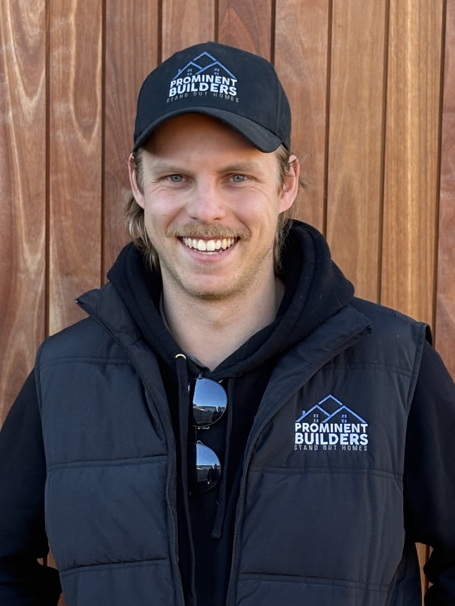 Joshua Lambert, Managing Director of Prominent Builders, in a branded cap and puffer vest against timber cladding