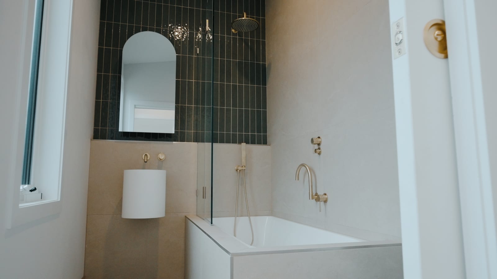 Bathroom with dark vertical tiling, an arched mirror, brushed gold tapware and a bath