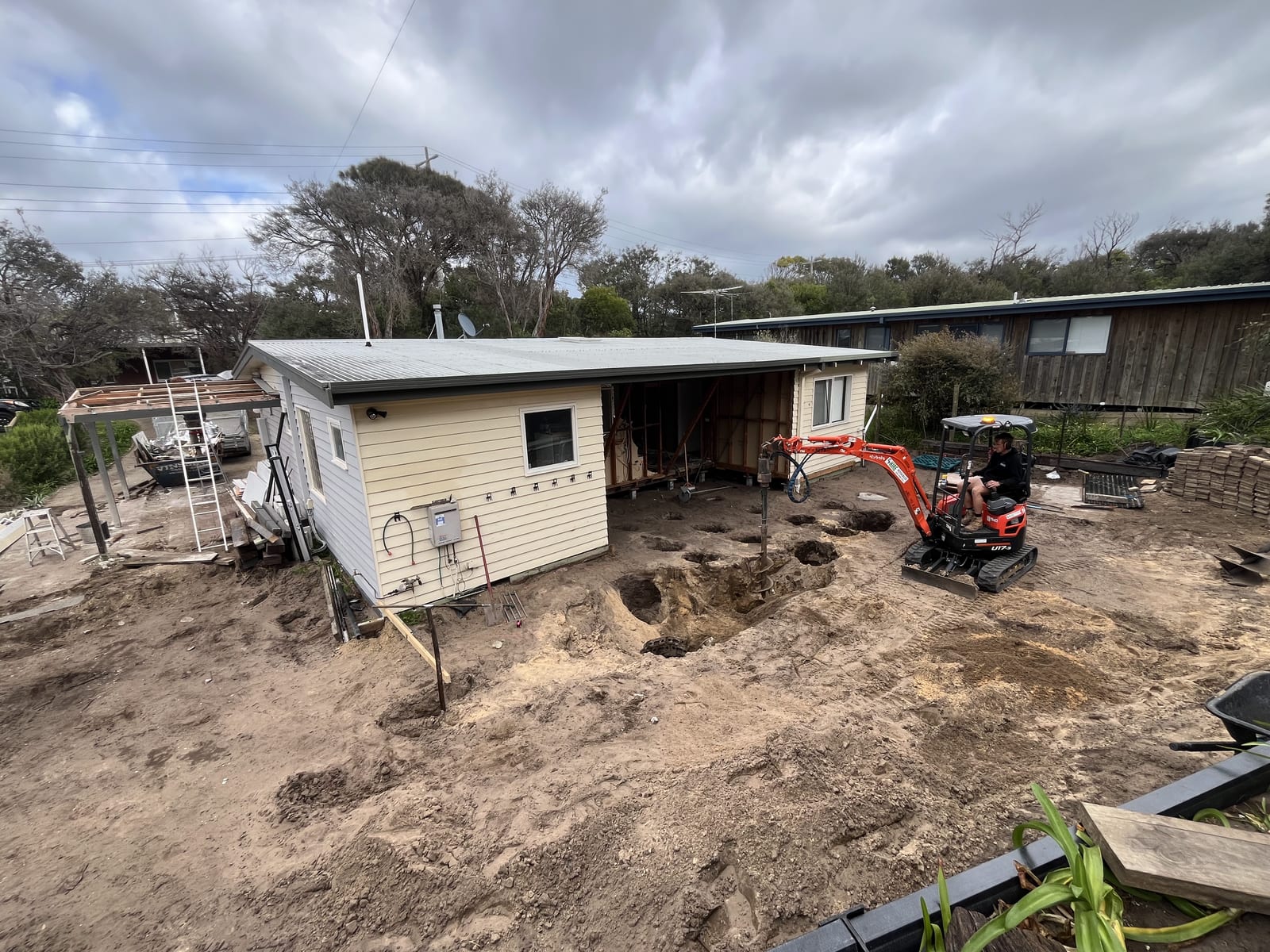 An excavator beside the original house with holes open in the ground where the footings are being dug