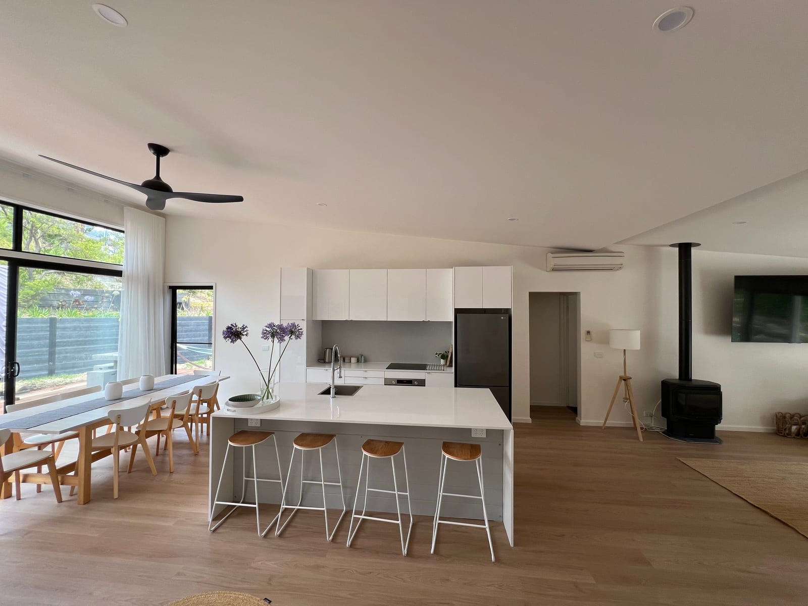 White kitchen with an island bench, timber stools and a long dining table under a black ceiling fan
