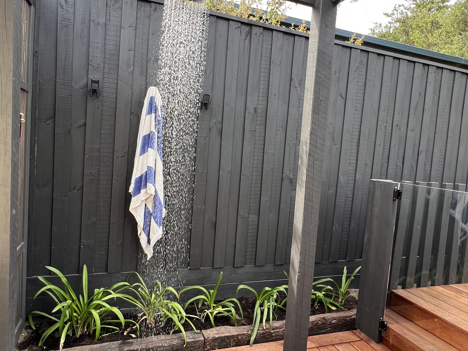 Outdoor shower running against a dark timber fence, mounted over hardwood decking with planting along the base