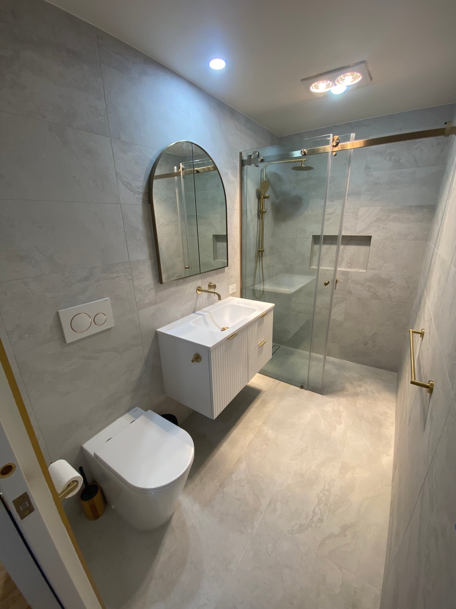The ensuite with a wall-hung toilet, fluted vanity, arched mirror and a gold-framed shower screen, tiled in the same pale 600 by 600 tile as the main bathroom