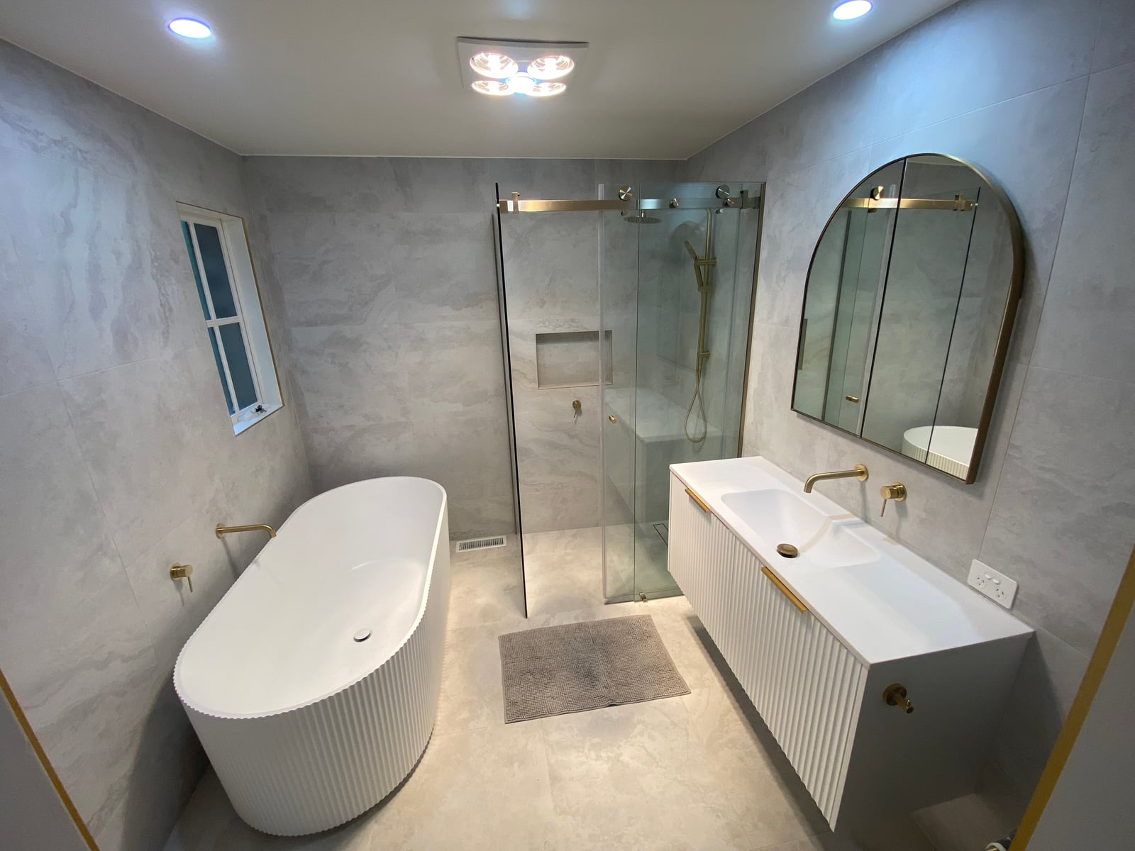 Completed Frankston main bathroom with a fluted freestanding bath, walk-in shower, fluted vanity and arched mirror, tiled floor to ceiling in pale 600 by 600 tile