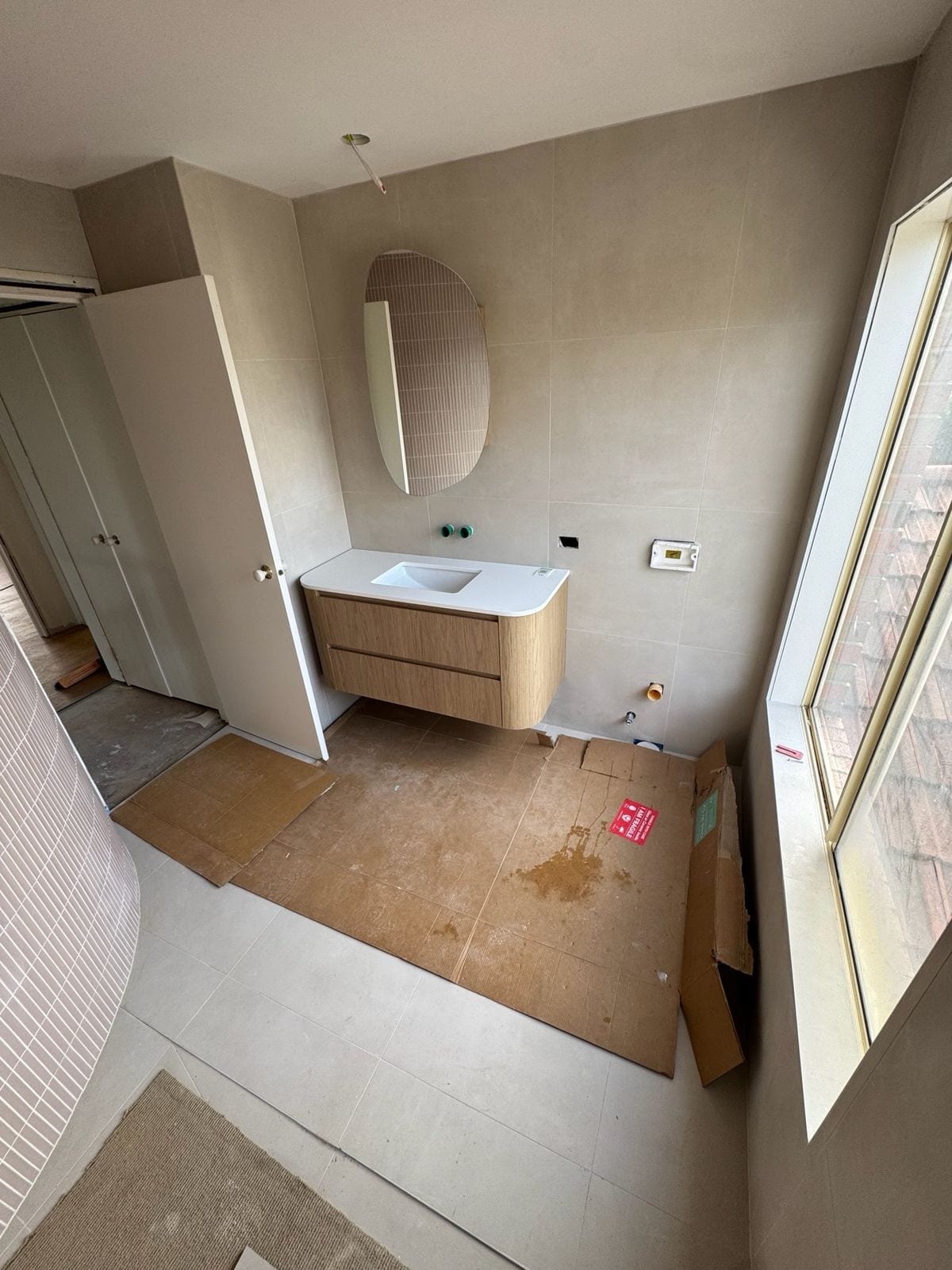 The upstairs main bathroom mid-fit-off: a wall-hung timber vanity with a curved end, an oval mirror, large pale wall tiles and a curved wall in narrow pink tiles on the left, with cardboard floor protection down
