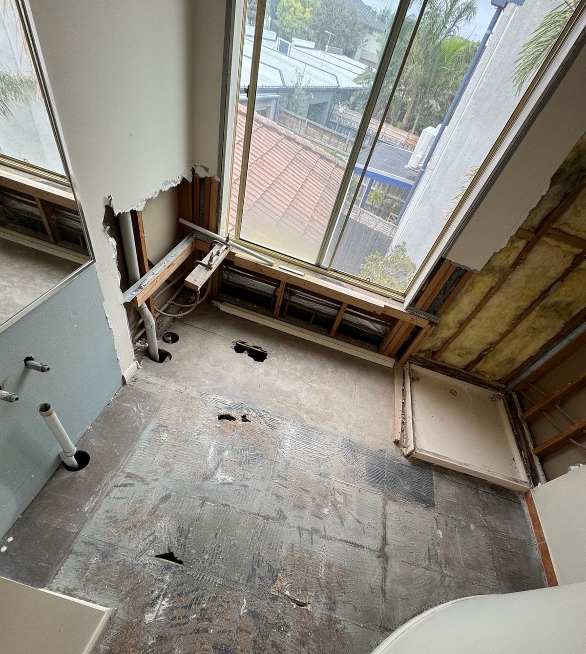 An upstairs wet area during strip-out: tiles and linings removed, wall frames and insulation exposed, the old shower base still in the corner and floor penetrations open beneath a large window