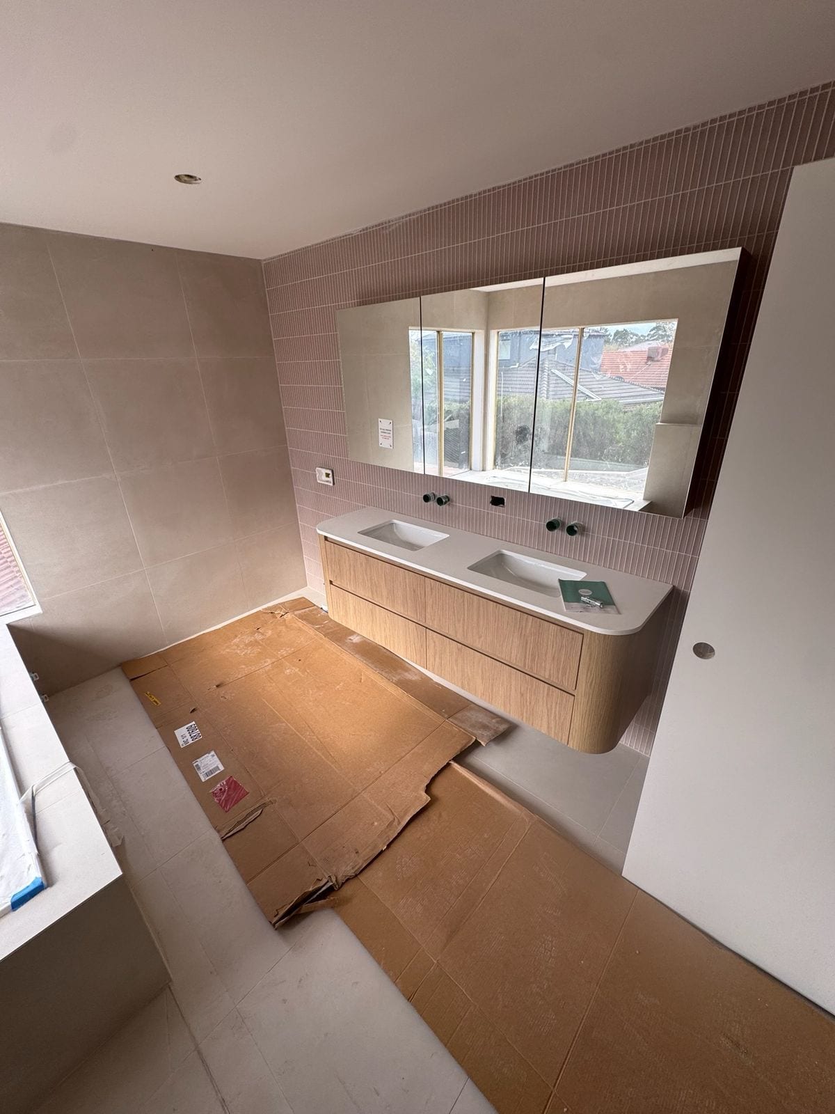 The new upstairs ensuite at Mordialloc mid-fit-off: a wall-hung timber vanity with a curved end and two undermount basins, a mirrored shaving cabinet, narrow vertical pink tiles behind the vanity and large pale tiles on the walls, with cardboard floor protection still down
