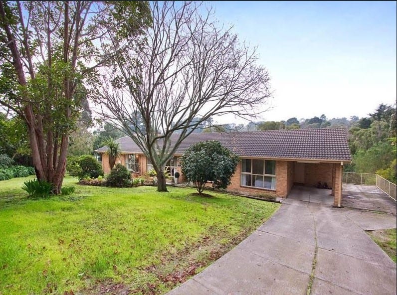 Single storey brick house at Mount Eliza with a tiled roof, set back behind a lawn and mature trees, with a concrete driveway running along the side to a small carport at the end of the building
