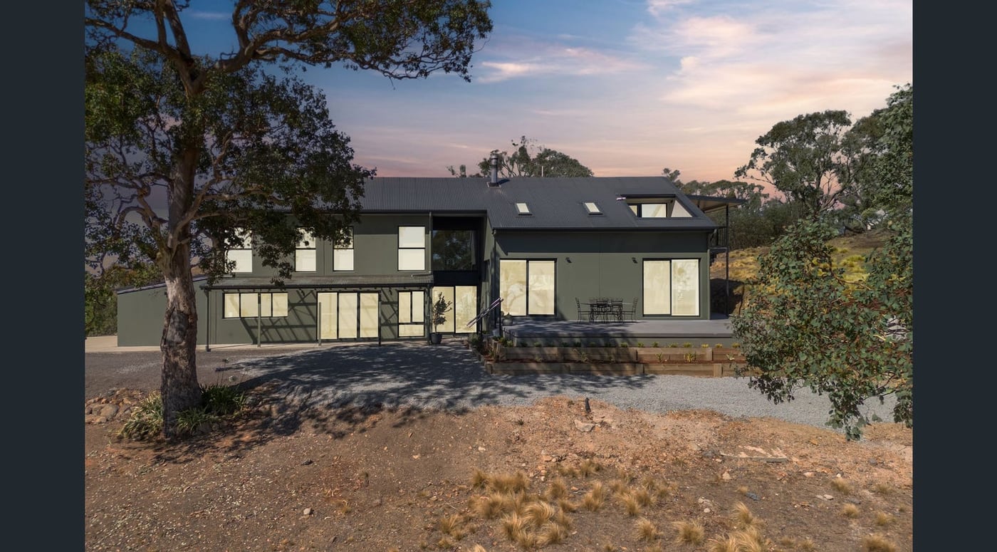 Two-storey precast concrete home in dark olive render at dusk, with a skillion metal roof, dormer windows and a raised deck, set among gum trees on a rural block