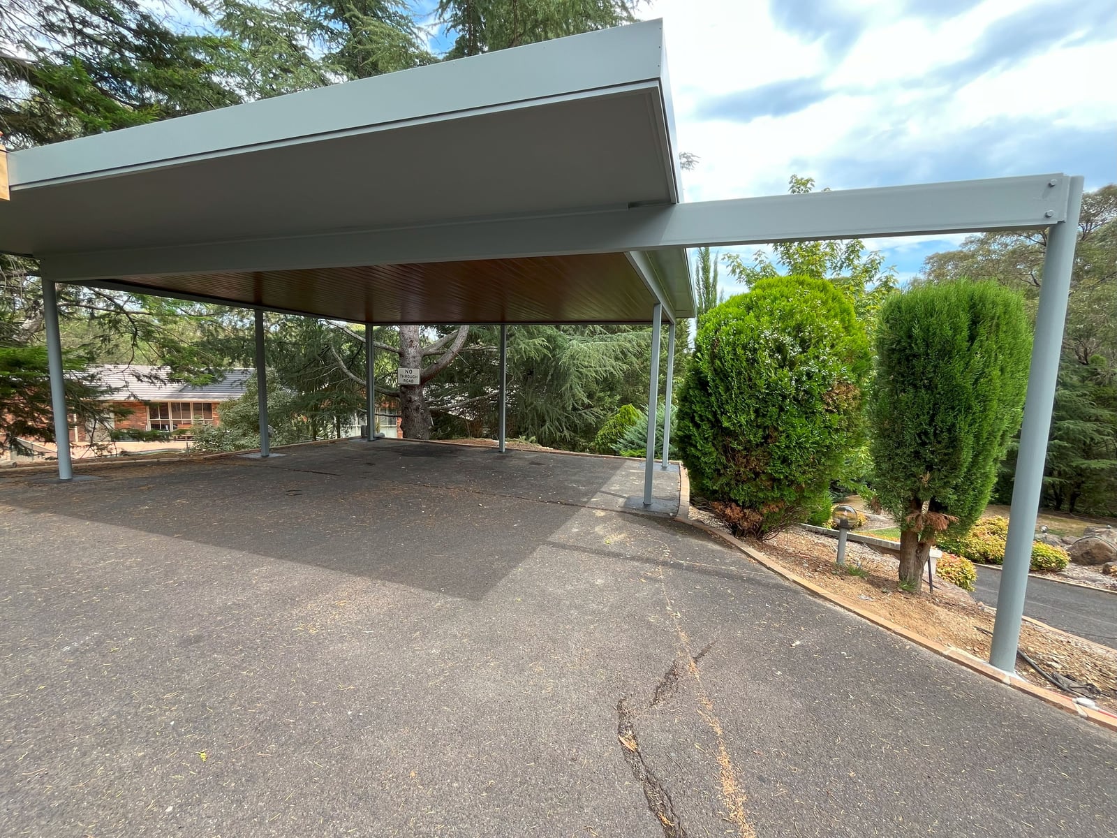 Completed freestanding carport with pale grey steel posts, a flat skillion roof and a timber-lined ceiling, over a driveway beside a landscaped garden